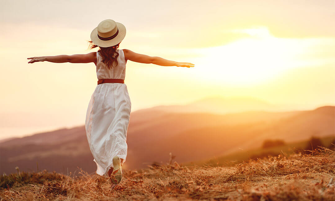 A woman wearing a brimmed hat with her arms spread wide running towards the sunset over a mountain.